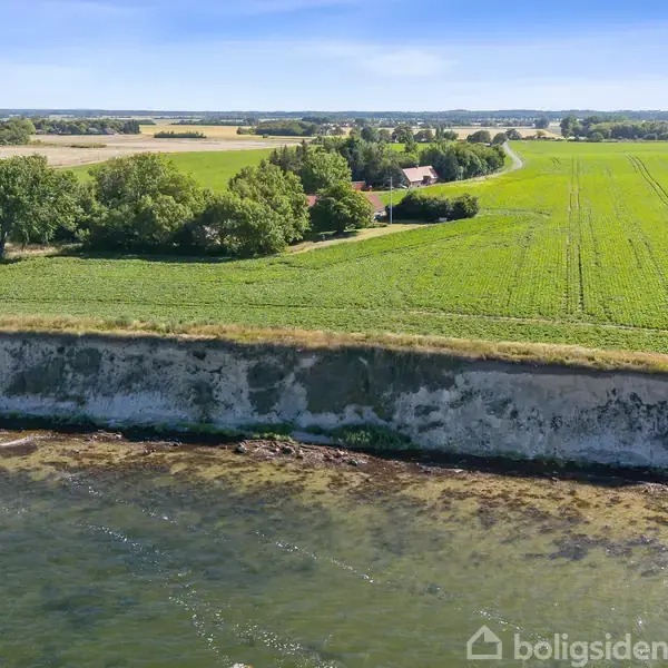 Vand møder en stejl klint i forgrunden, hvorpå en stor mark strækker sig mod horisonten. En vindmølle står stille blandt træer. Boligsiden.
