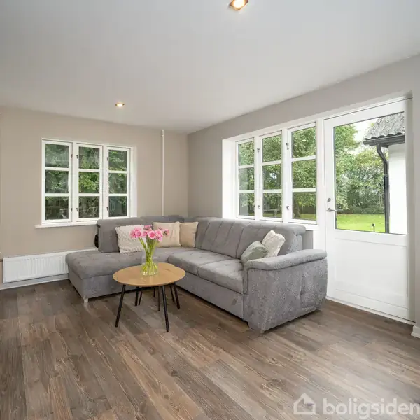 Grå sofa står i en stue med trægulv. Sofaen har puder og er ved et rundt bord med blomster. Store vinduer og dør fører ud til en grøn have.