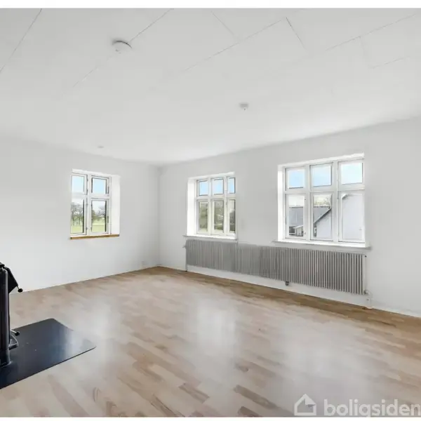 Et tomt, lyst rum med trægulv har en sort brændeovn i hjørnet. Store vinduer på to vægge giver naturligt lys. Væggene er hvide og en radiator ses under vinduet.