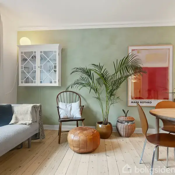 En stue med en grøn væg, en grå sofa til venstre, planter i midten, et rundt træbord til højre, og et Mark Rothko kunstplakat i baggrunden.