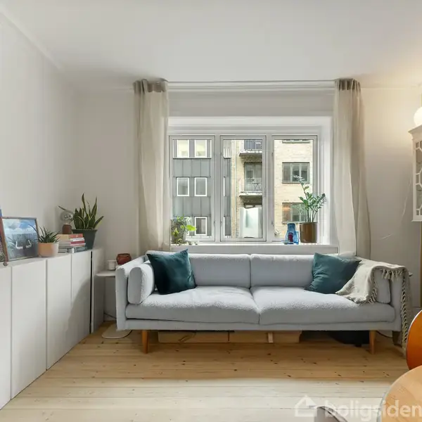 Grå sofa står ved vindue med grønne puder, omgivet af hvide skabe og planter. Kunstværker er placeret på skabene i en lys stue med trægulv og gardiner.