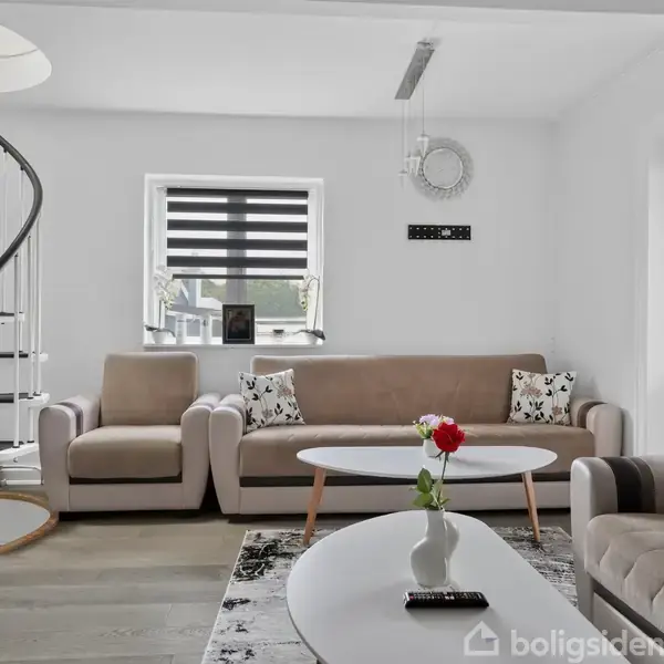 En stue med en beige sofa og sofabord, udsmykket med blomstervase. En spiraltrappe står til venstre. Vinduet bagved har mørklægningsgardin. Diverse pyntepuder er på sofaerne.