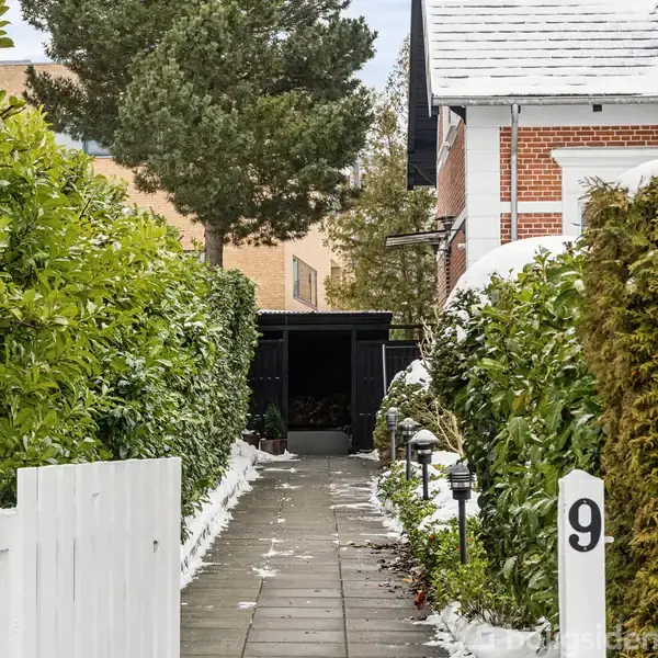 En sti fører op til en åben skur fyldt med brænde. Omgivet af hække med snedækkede grene, står der en hvid port med tallet "9" til højre.