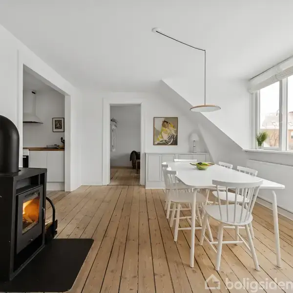 En brændeovn står i et lyst spisestueområde med trægulv. Et hvidt spisebord med seks stole er placeret ved et stort vindue. Maleri hænger på væggen.