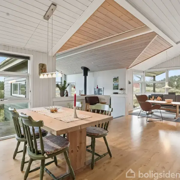 Spisestue med træbord og fire stole, hvor et stearinlys er placeret. I baggrunden ses et moderne opholdsområde med sofaer. Store vinduer giver udsigt til naturen udenfor.