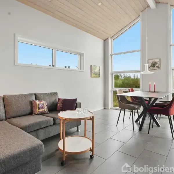 Sofa med farverige puder står ved siden af et lille rundt bord på hjul i lys stue. Store vinduer bagved giver udsigt til en grøn have.