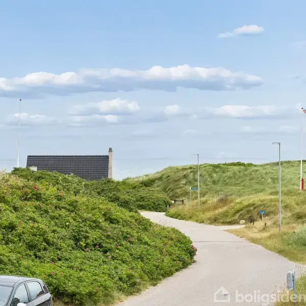Et hvidt hus med mørkt tag ligger i et grønt område nær kysten, omgivet af andre boliger og veje, med havet i baggrunden under en skyet himmel.