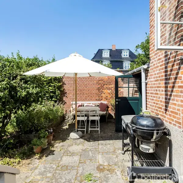 Haveområde med betonfliser og en grill. Et bord med stole står under en stor parasol. Murstensbygning og grønne buske omgiver terrassen.