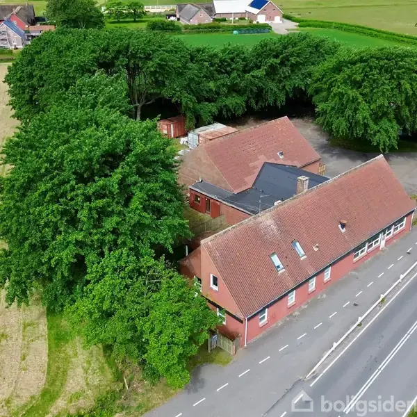 Rødt hus med rødt tag ligger ved en vej omgivet af tætte, grønne træer. En mark ses til venstre, og vejen løber parallelt på højre side. Ingen synlig tekst.