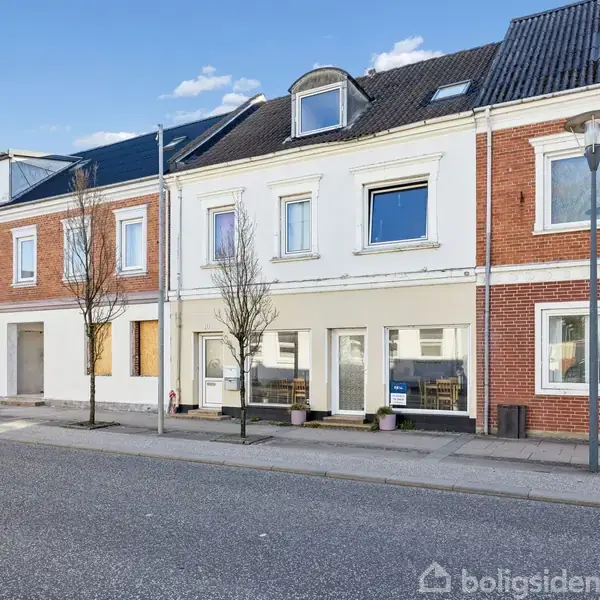 Rækkehus med røde og hvide facader står roligt langs en ren gade. Foran huset er der træer og gadelamper. Skyfri blå himmel danner en fredelig baggrund.