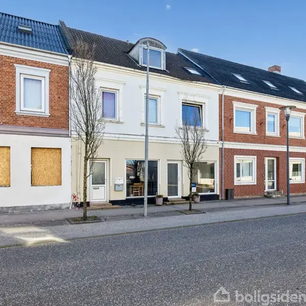 Bygning med flere etager står langs en gade. Flere vinduer på facaden er synlige. Omgivet af asfalteret vej og enkelte træer. En bygning har tilplankede vinduer.