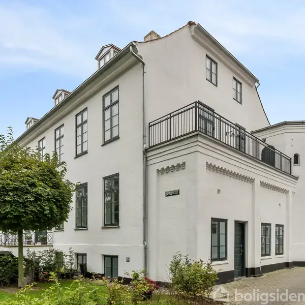Hvid ejendom med sort-rammende vinduer og en altan i øverste etage. Huset står i en grøn have med buske og træer, omgivet af en klar blå himmel.