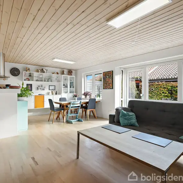 Grå sofa med grønne puder står i en moderne stue. Store vinduer lader lys ind. Spisebord med stole i baggrunden. Køkken til venstre med hylder og udsmykning.