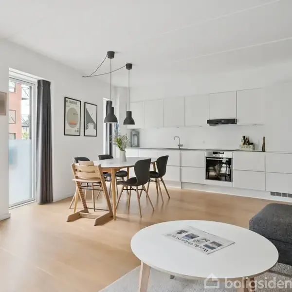En moderne stue-køkken-alrum med hvidt køkken, spisebord og stole. Store vinduer med mørke gardiner lyser rummet op. En grå sofa og hvidt sofabord står i forgrunden.