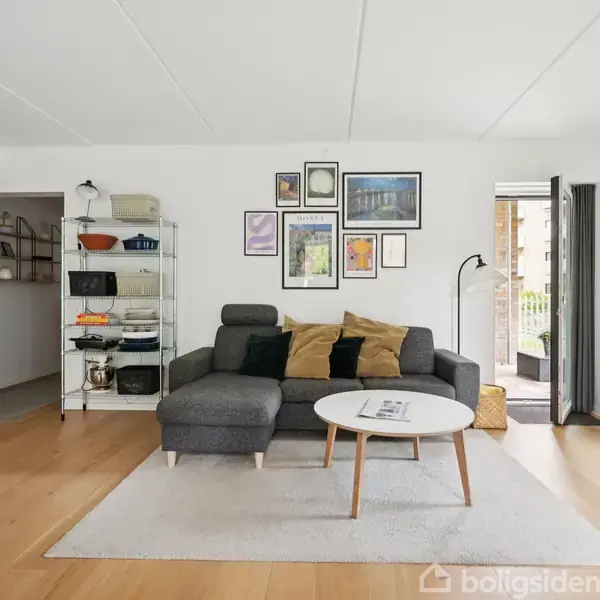 En grå sofa står i en stue, pyntet med puder. En væg har kunstbilleder. Et sofabord står foran sofaen. Bagved er en reol med køkkengrej, og en dør fører ud til en balkon.