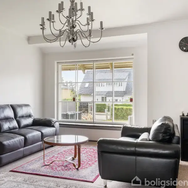 Sort lædersofa står ved siden af et lille rundt bord på tæppe. En lysekrone hænger fra loftet. Store vinduer viser udsigt til have og hus. Klokke på væggen.