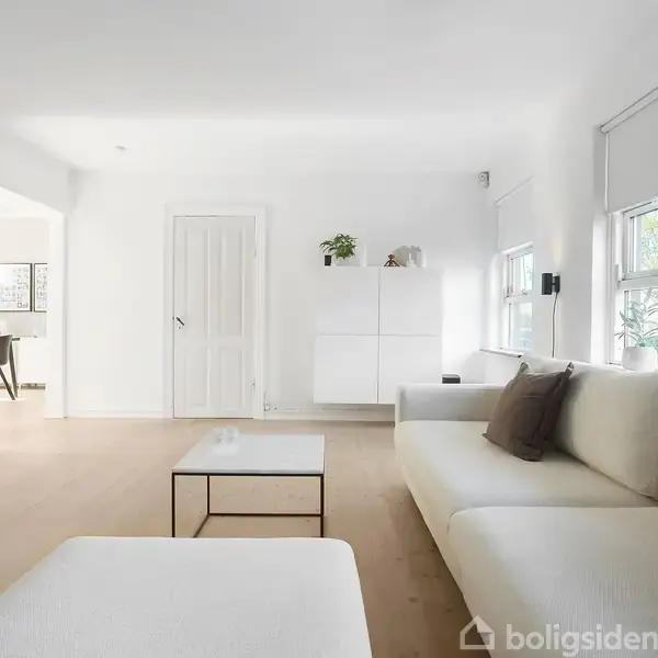 En lys stue med en hvid moderne sofa og sofabord ved et stort vindue. Væggene er hvide med minimalistisk kunst, og en dør fører til et møbleret rum.
