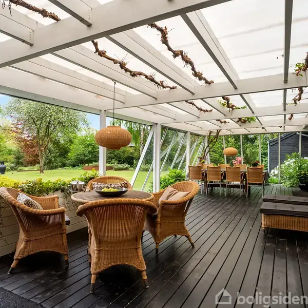 Fletmøbler står på en overdækket terrasse med sort trædæk, omgivet af græsplæne og frodig have. Terrasseoverdækningen er dekoreret med grene, hvilket skaber en hyggelig og naturlig atmosfære.