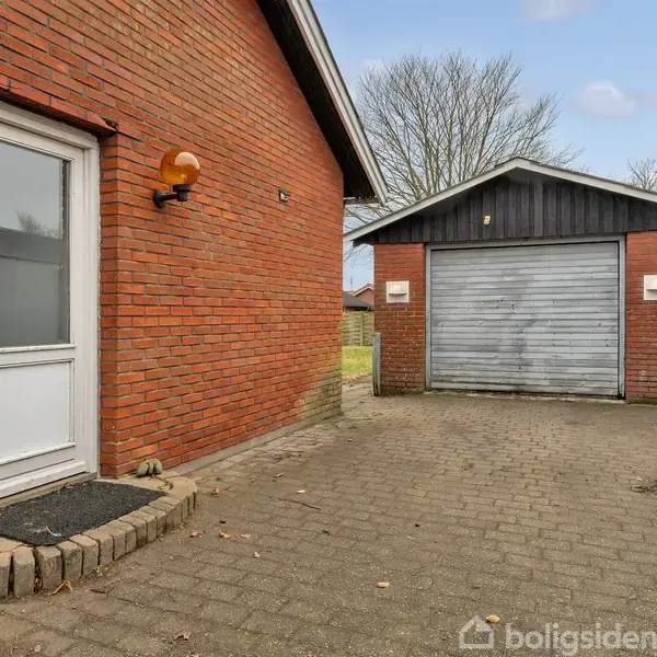 Rød murstensbygning med en hvid dør og sort garage i baggrunden med metalskydedør. Belægning af grå sten danner gårdspladsen. Træer er synlige i baggrunden under en overskyet himmel.