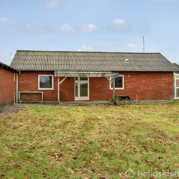 En murstenshus står stille i en have. Græsplænen er dækket af visne blade. I baggrunden ses himlen med få skyer, og til venstre er et træ synligt.