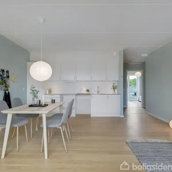Et lyst og moderne køkken-alrum med et spisebord og fire grå stole. Køkkenet har hvide skabe, og der er en rund lampe hængende fra loftet. En smal gang ses i baggrunden.