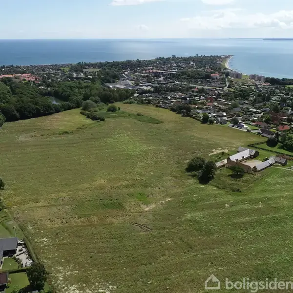 En stor, åben mark strækker sig mod en kystby. Markerne er grønne og omkranset af spredte huse. I baggrunden ses en kystlinje og hav.
