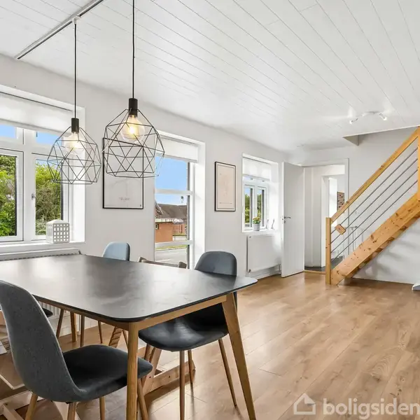 Et moderne spisebord med seks stole står i et lyst rum. To geometriske lamper hænger over bordet. Trætrapper fører opad i rummet med trægulv og store vinduer.