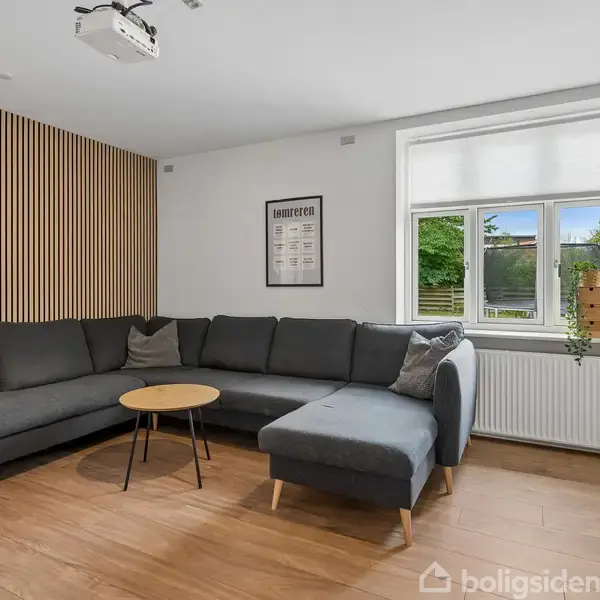 En mørkegrå hjørnesofa står i en stue med trægulv, flankeret af et lille rundt bord. En plakat hænger på væggen, og et vindue giver udsigt til en have.