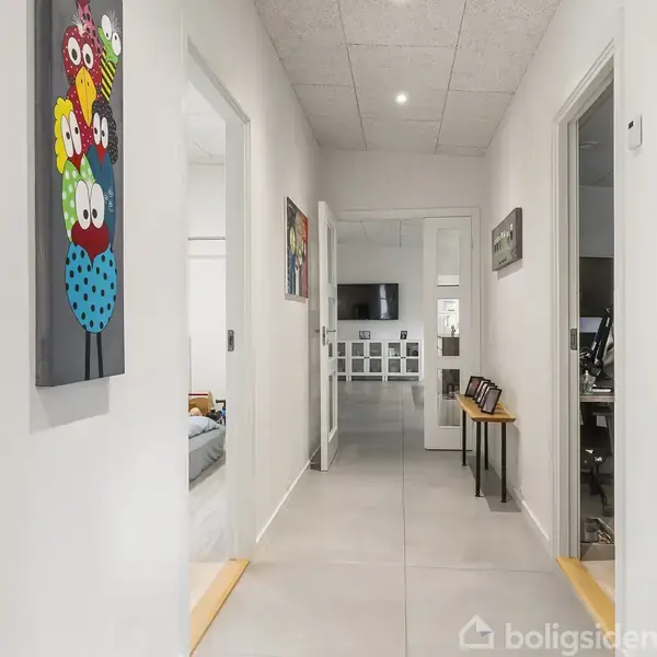 En lang gang med lyse vægge har kunst på væggen til venstre. En dør i slutningen åbner mod et lyst rum med TV og billeder på væggen.