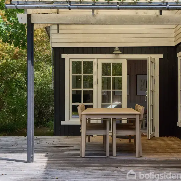 En træterrasse med et spisebord og stole stående under en pergola. Terrassen er foran et sort træhus med hvide vinduer og dør, omgivet af grønne buske og træer.