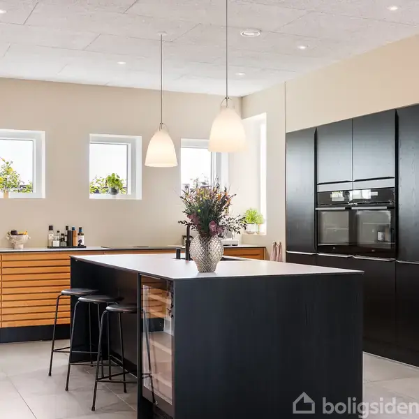 Et moderne køkken med en mørk ø med barstole. Ovenpå er der hængende lamper og blomster i en vase. I baggrunden ses skabe, vinduer med planter og køkkenredskaber.