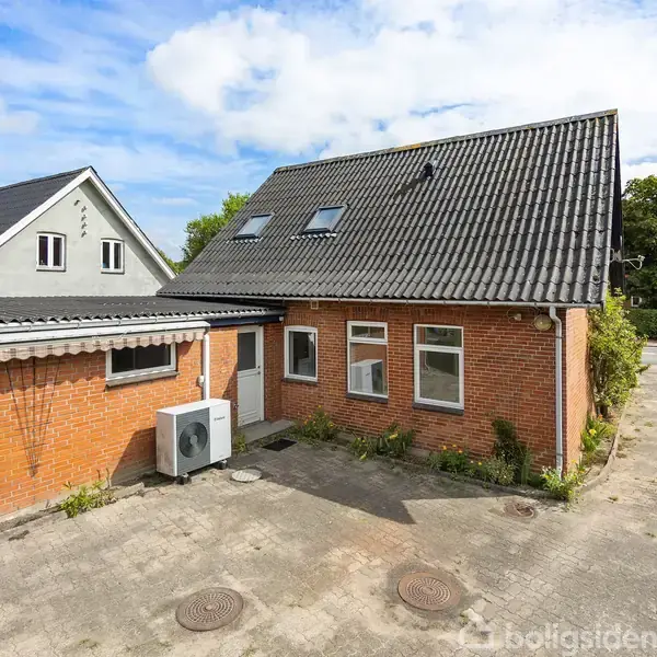 Rødt murstenshus med sort tag, flere vinduer og en hvid dør. En airconditionenhed er monteret på facaden. Omgivet af en flisebelagt gårdsplads og grønne områder.