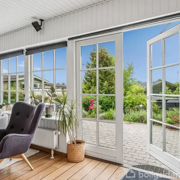 Store glasdøre åbner ud mod en terrasse med brosten og frodig have. Indenfor står komfortable stole på trægulv ved siden af planter og store vinduer, der indrammer udsigten.