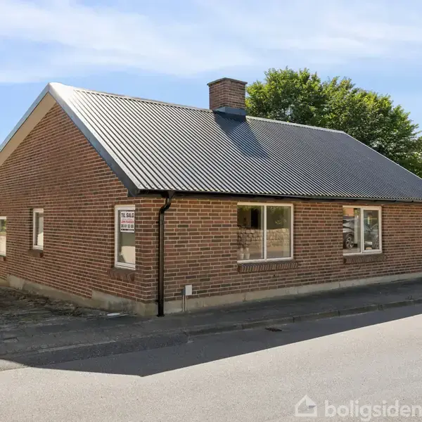 En muret en-etagers bygning med gråt tag står ved en vej. I baggrunden ses grønne træer og græsarealer. Der er et skilt ved siden af huset, som lyder: "TIL SALG, Jørgen Møller & Petersen, Ejendomsmægler."