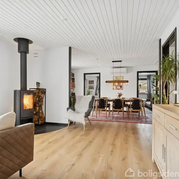 Brændeovn står tændt i lys stue med træmøbler, åbent til spiseplads med stort bord og stole. Store vinduer slipper naturligt lys ind på trægulvet og dekorativt tæppe.