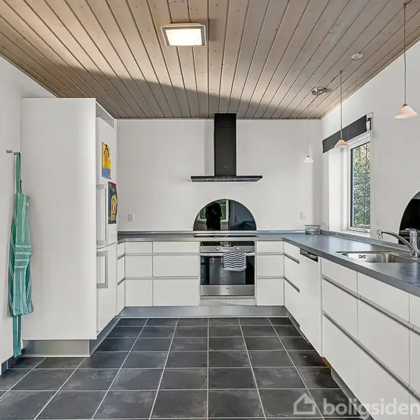 Et lyst køkken med hvide skabe og sorte fliser. En ovn og emhætte i midten. Hængende grønne forklæder til venstre. Vinduer og lamper giver naturligt lys.