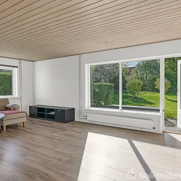 Stue med sofa og sofabord ved vindue, der åbner ud til en grøn have. Trægulv og træloft skaber en lys og indbydende atmosfære. Ingen tekst er synlig.