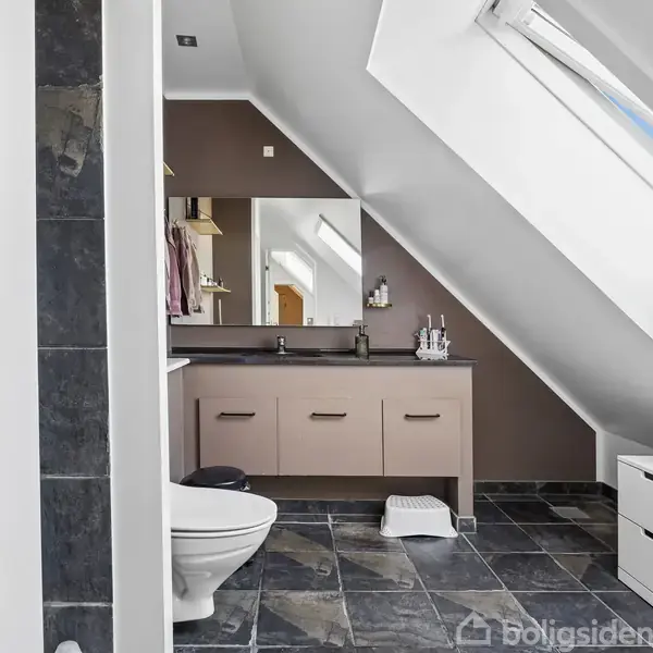 Badeværelse med toilet og håndvask under skråvæg. Stor spejl over vasken reflekterer dagslys fra ovenlysvindue. Flisegulv og vægpaneler i grå nuancer.