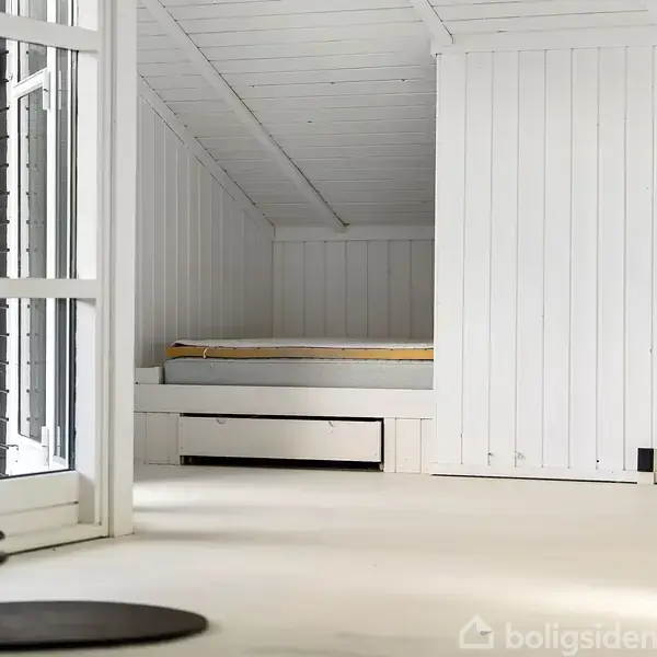 En hvidmalet loftsrum med en enkeltseng indbygget i væggen. Rummet har skråloft og er delvist synligt gennem en åben dør. Ingen tekst er til stede.