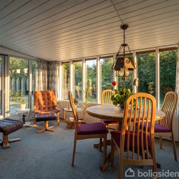 En spisestue med et rundt bord og træstole med lilla hynder. En lænestol står ved siden af, med glasvinduer i baggrunden, der viser en terrasse og grønne træer udenfor.