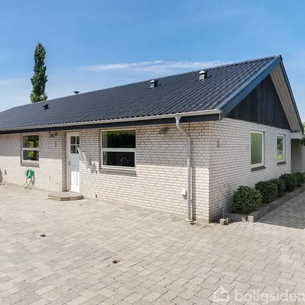 En enkeltplans murstensvilla med sort tag står på en stor brolagt gårdsplads. Omgivet af træer og buske under en klar blå himmel.