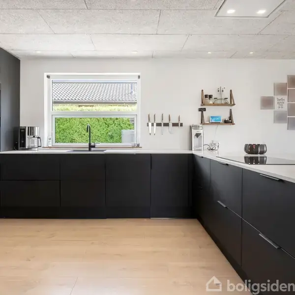 Et moderne køkken med sorte skabe og en hvid bordplade strækker sig langs væggen. Et stort vindue giver udsyn til en grøn hæk, mens lyse trægulve kompletterer indretningen.