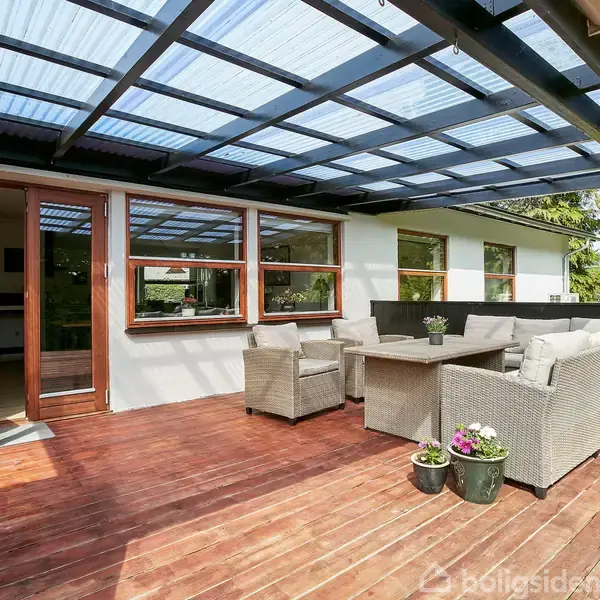 Terrasse med flettede møbler står under et halvtag af gennemsigtigt materiale. Møblerne inkluderer en sofa, to stole og et bord. Planter i potter tilføjer et dekorativt touch.