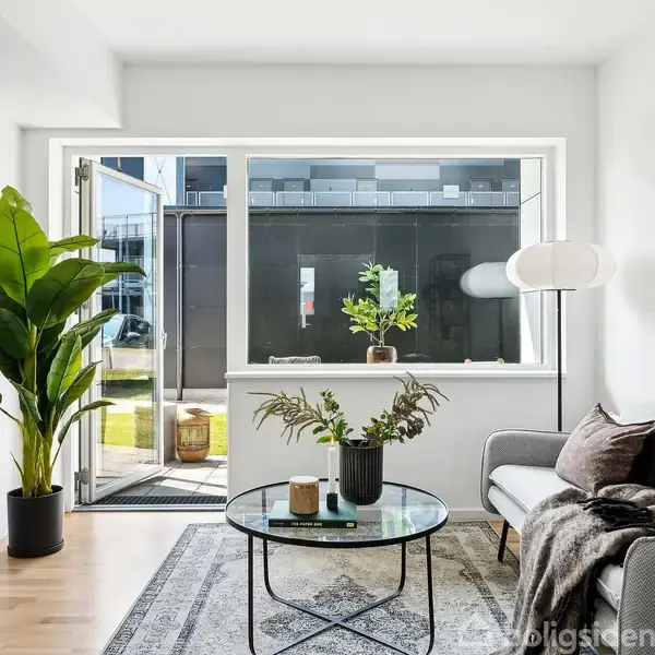 Stue med grå sofa, rundt glasbord med planter og lysestage. Stor tropical plante ved siden af en reol. Store vinduer åbne til en terrasse med udsigt til bygninger.