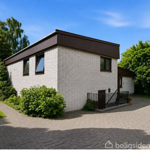 Hvid murstenshus med skråt tag ligger til højre på billedet. Foran huset er en flisebelagt indkørsel. Grønne buske og træer omgiver området under en klar blå himmel.