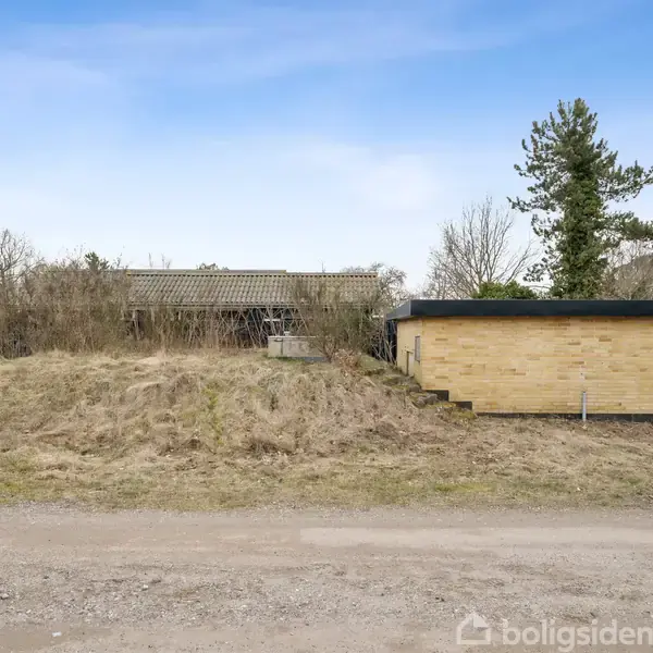 En lille bygning med fladt tag står ved siden af en jordhøj. Omkring er der træer og buske, og forbi løber en grusvej. Skyfri, lys himmel i baggrunden.