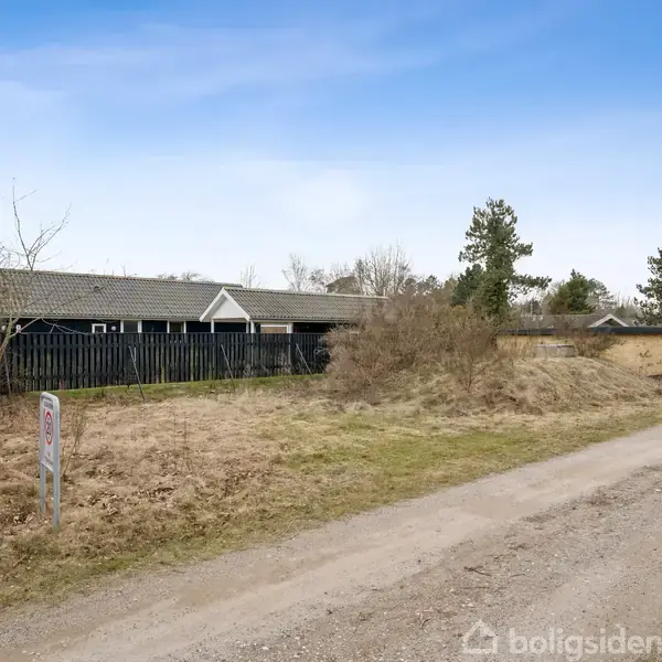 En lav bygning med mørke træfacader står på en græsmark omgivet af buske og træer. En grusvej løber ved siden af, og et skilt står nær vejen.
