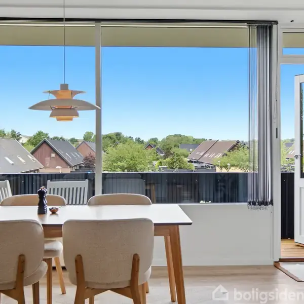 Spisebord med fire stole, beliggende i en lys spisestue. Store vinduer åbner ud mod en altan, der viser et beboelsesområde med træer og huse i baggrunden.