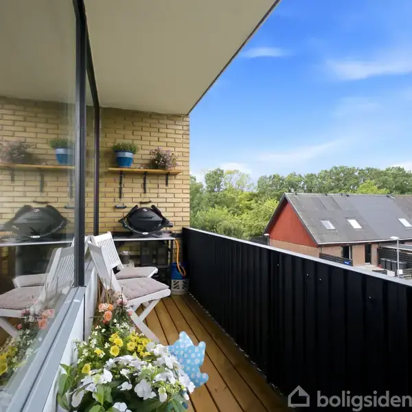 Altan med træmøbler og blomster i potter. Grill står mod en murstensvæg. Omkringliggende bygninger ses i baggrunden, og en blå himmel er over området.