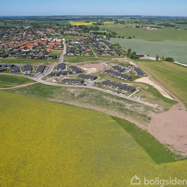 Bebyggelse med rækkehuse ligger på en bakketop. Omkringliggende marker og åbent land strækker sig til horisonten. Byområder er synlige i baggrunden. Marker viser gule blomstrende afgrøder.
